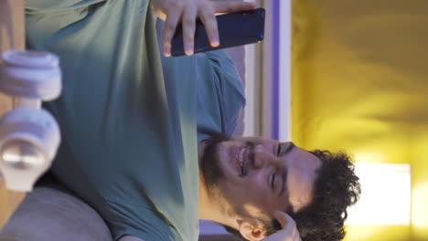 Vertical-video-of-Man-lying-on-the-sofa-at-night-texting-on-the-phone.-Happy-and-in-good-spirits.
